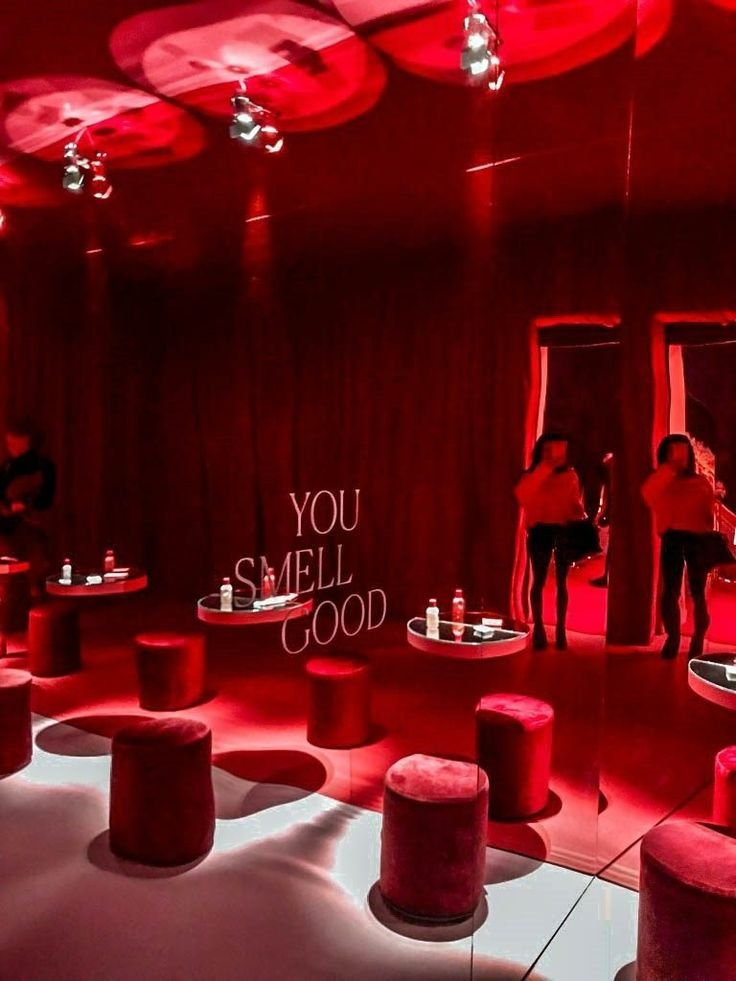 A red-themed room with round seating stools and small tables, with bottles and glasses on the tables. A mirrored wall reflecting two women taking photos, and the phrase 'You Smell Good' written on the wall.