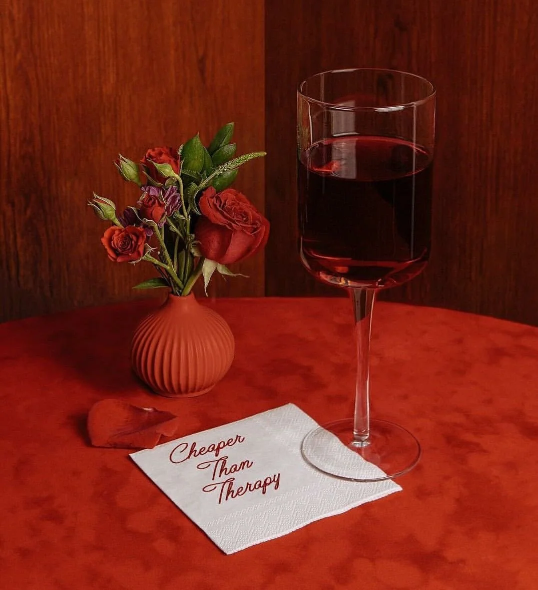 A red-tiled table with a small vase of red roses, a glass of red wine, a white napkin with red text that reads 'Cheaper Than Therapy,' and a single rose petal.