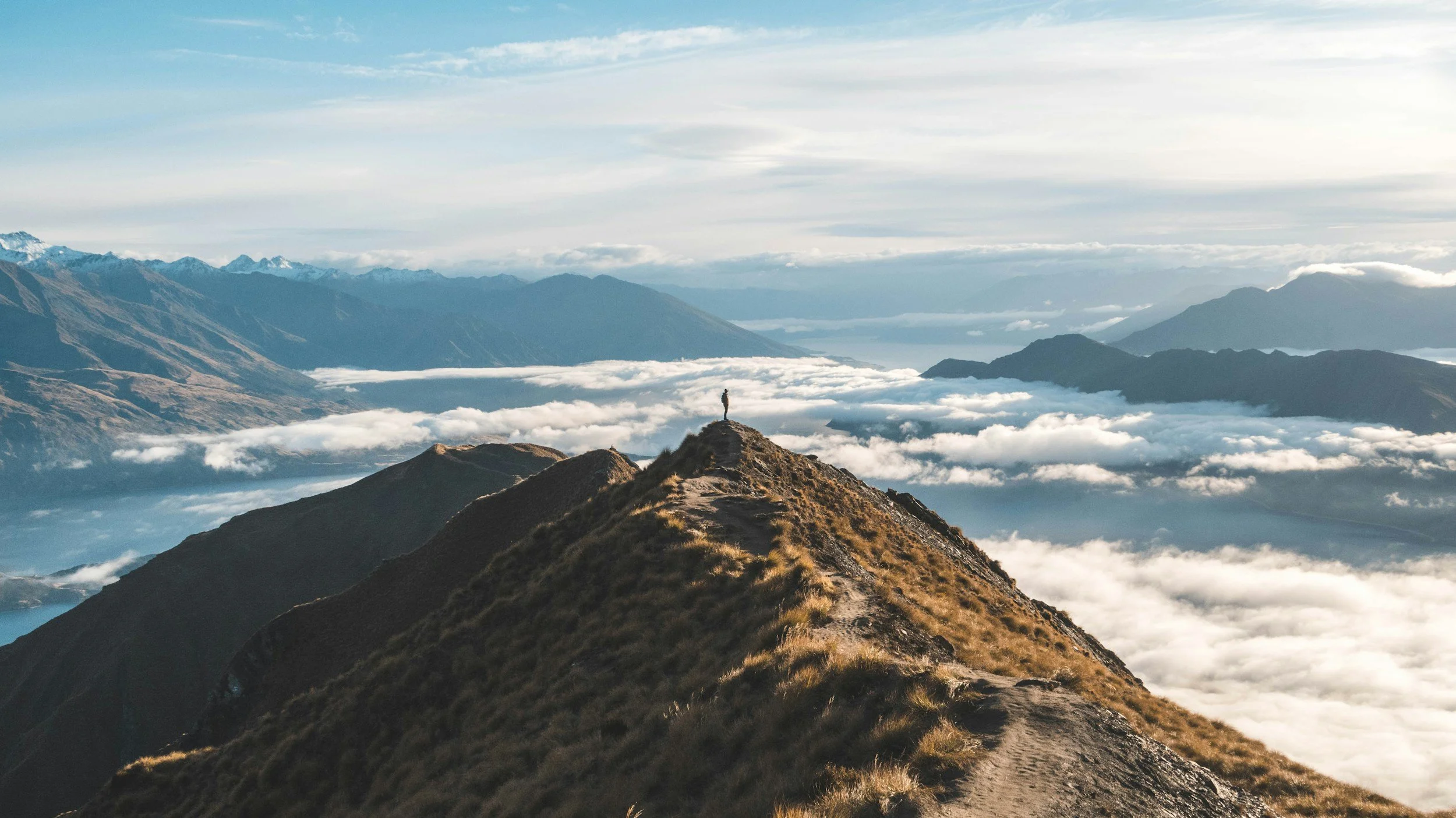 A person standing on a mountain ridge overlooking a sea of clouds and distant mountain ranges shows he is dreaming about AI but needs collaboration to materialize the dream.