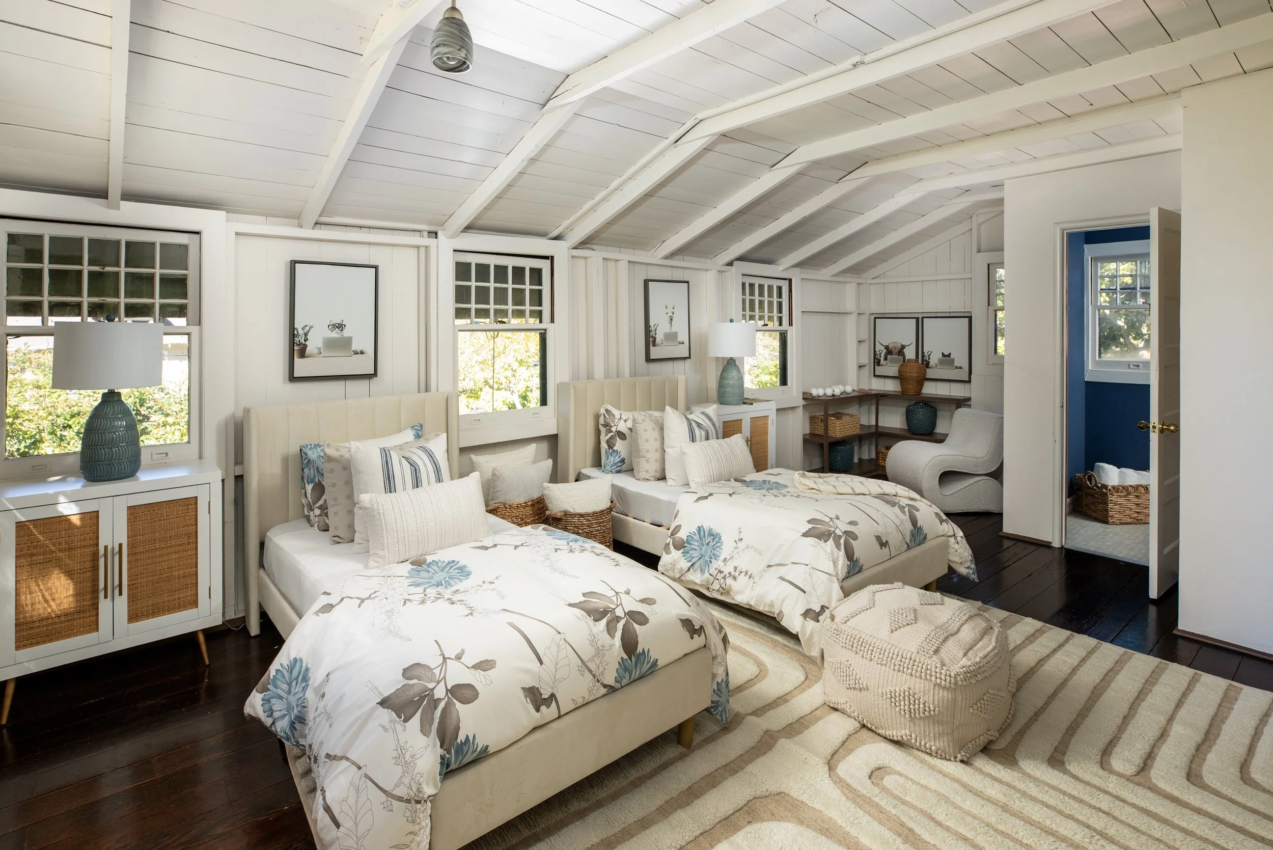 A bright bedroom with two twin beds with floral bedding, a beige armchair, and a white ceiling with exposed beams. There are windows with views of green trees, and the room has wooden flooring and decorative pillows and lamps.