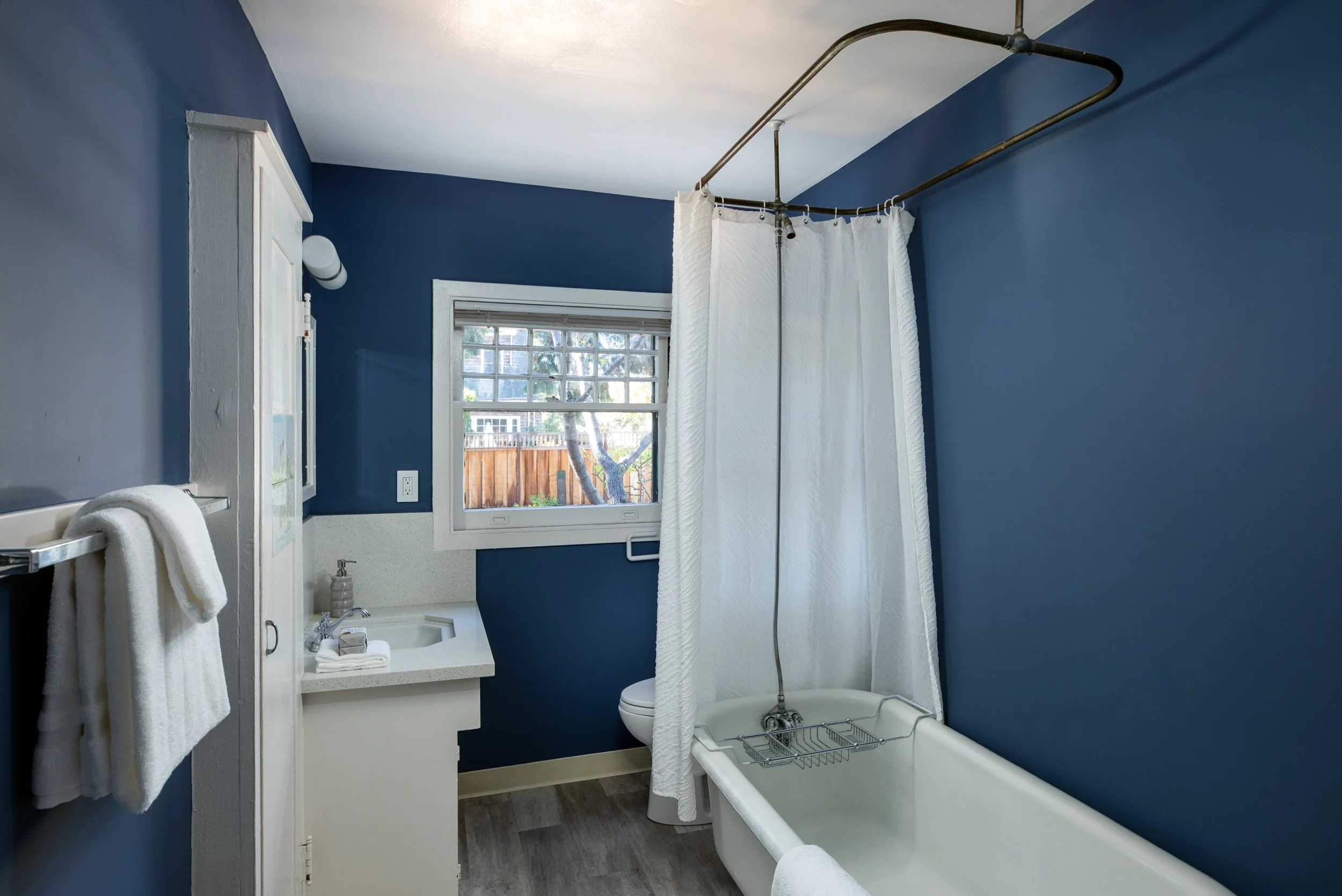 Bathroom with blue walls, white bathtub with shower curtain, white sink with soap dispenser, window showing outdoor trees and fence, towel hanging on rack, and small white cabinet.