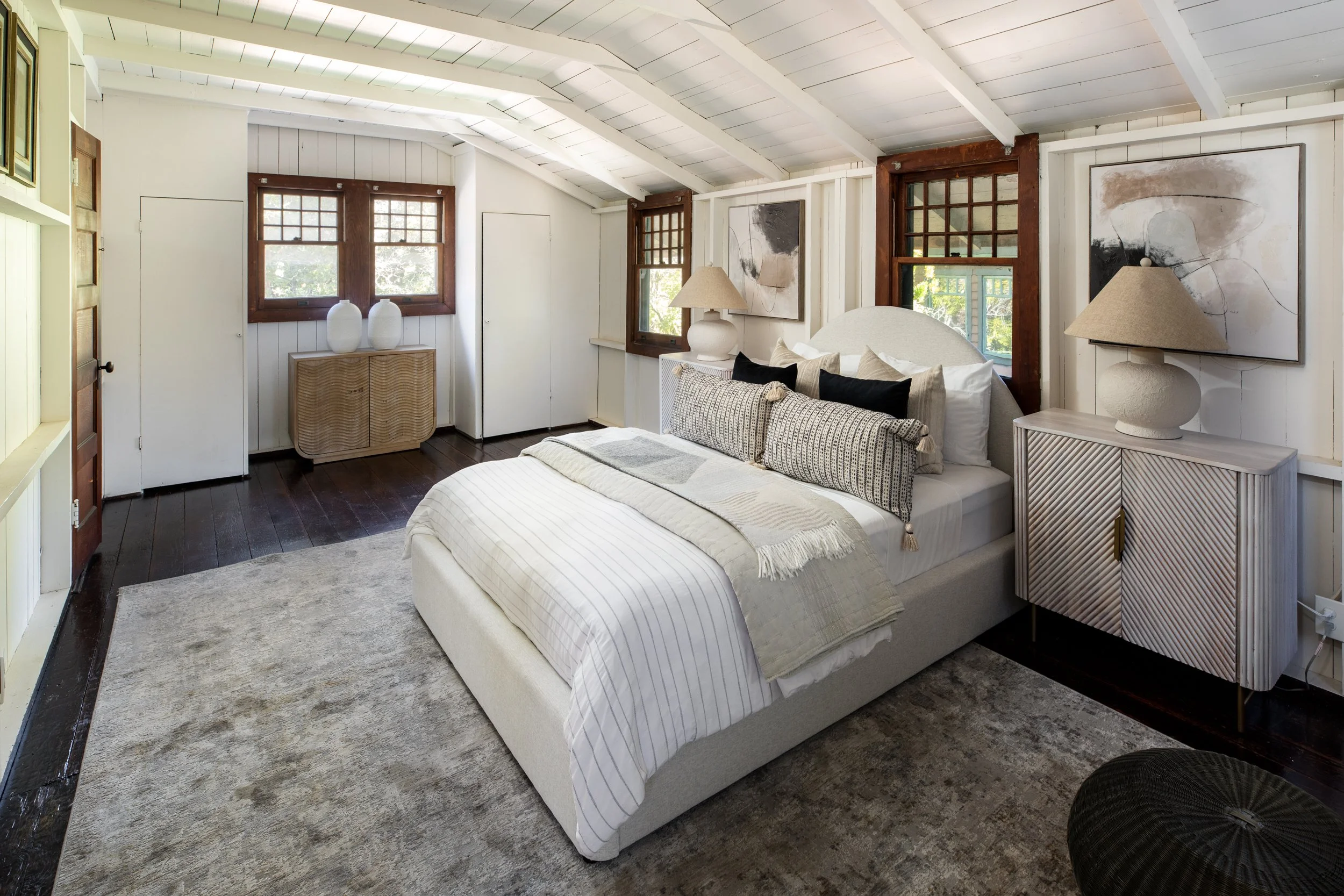 A cozy bedroom with a white bed, dark hardwood floors, and white walls with windows. There are lamps, artwork, and decorative pillows and blankets on the bed.