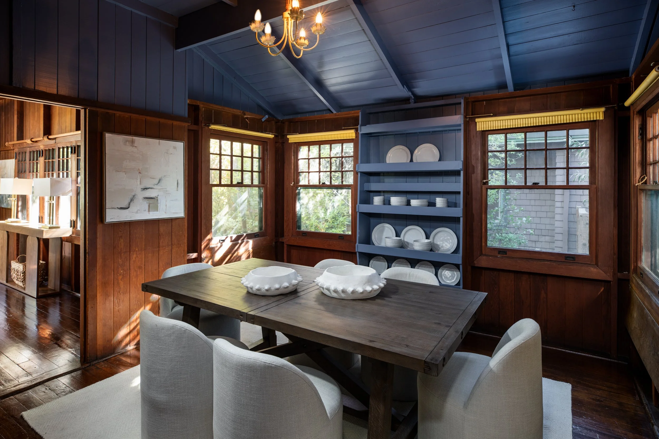 Dining room with a wooden table, six white chairs, blue walls, windowed walls, and a blue built-in china cabinet filled with white dishes, chandelier overhead, and sunlight coming through the windows.