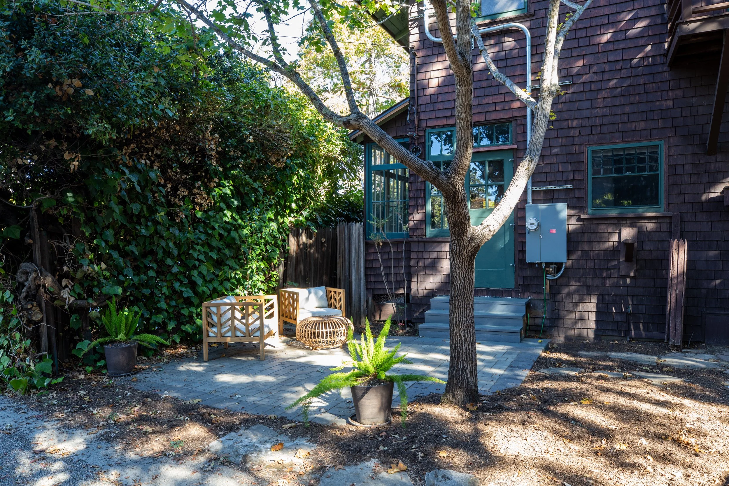 Small outdoor patio area with two wooden chairs, a round wicker table, and potted plants, next to a tree and the back of a dark wooden house with steps leading to a green door.