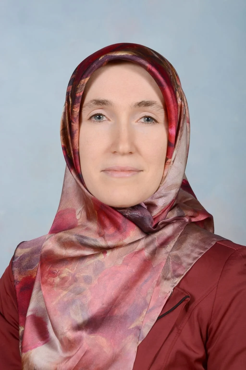 A woman with fair skin and light blue eyes, wearing a floral patterned headscarf in shades of pink, red, and purple, and a maroon jacket, facing and looking directly at the camera with a neutral expression.