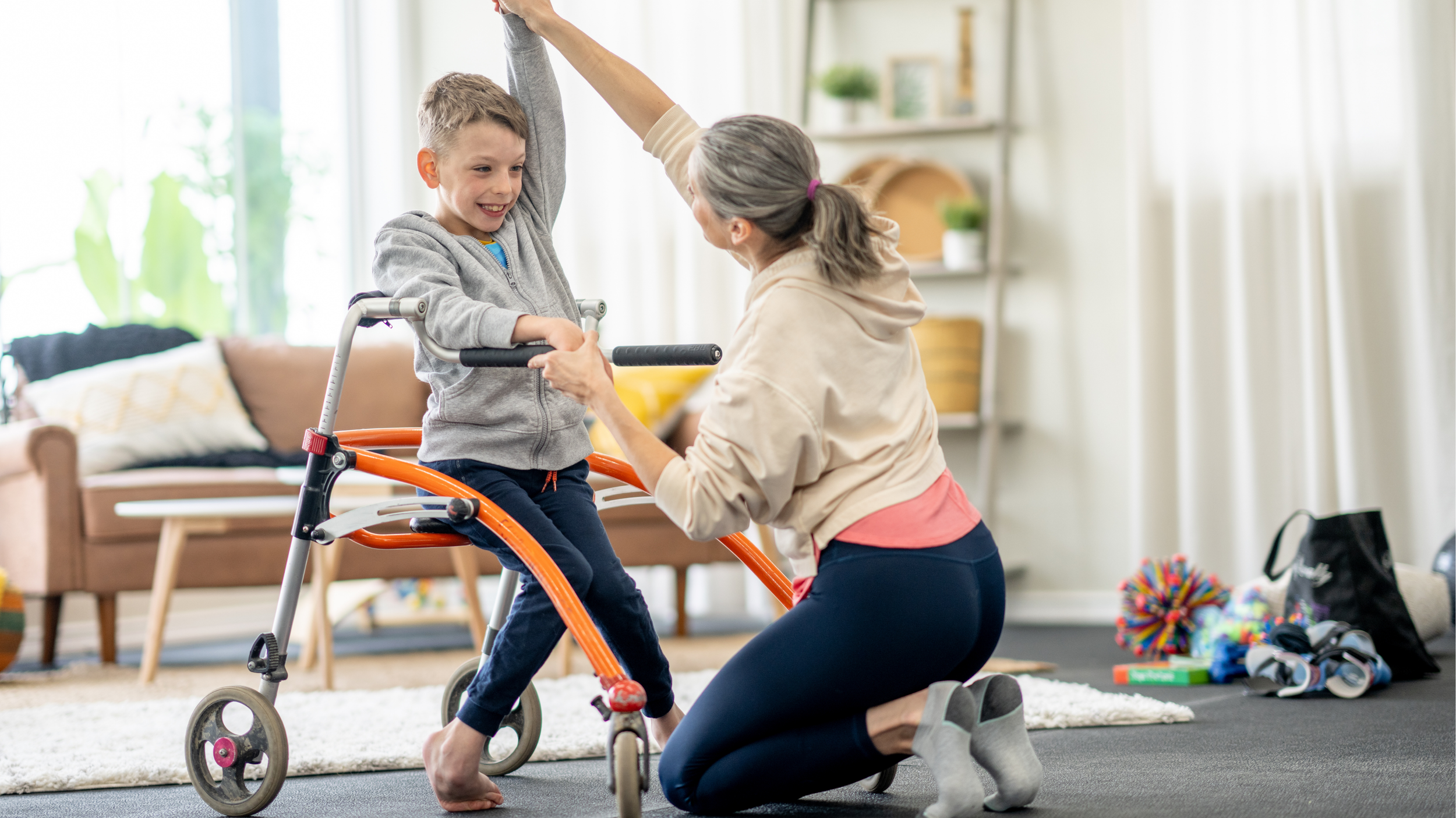 An occupational therapist  assisting a boy, who is standing on a walker, inside a living room with natural light. The occupational therapist is kneeling on the carpet, holding the boy's hand and arm, helping him balance.