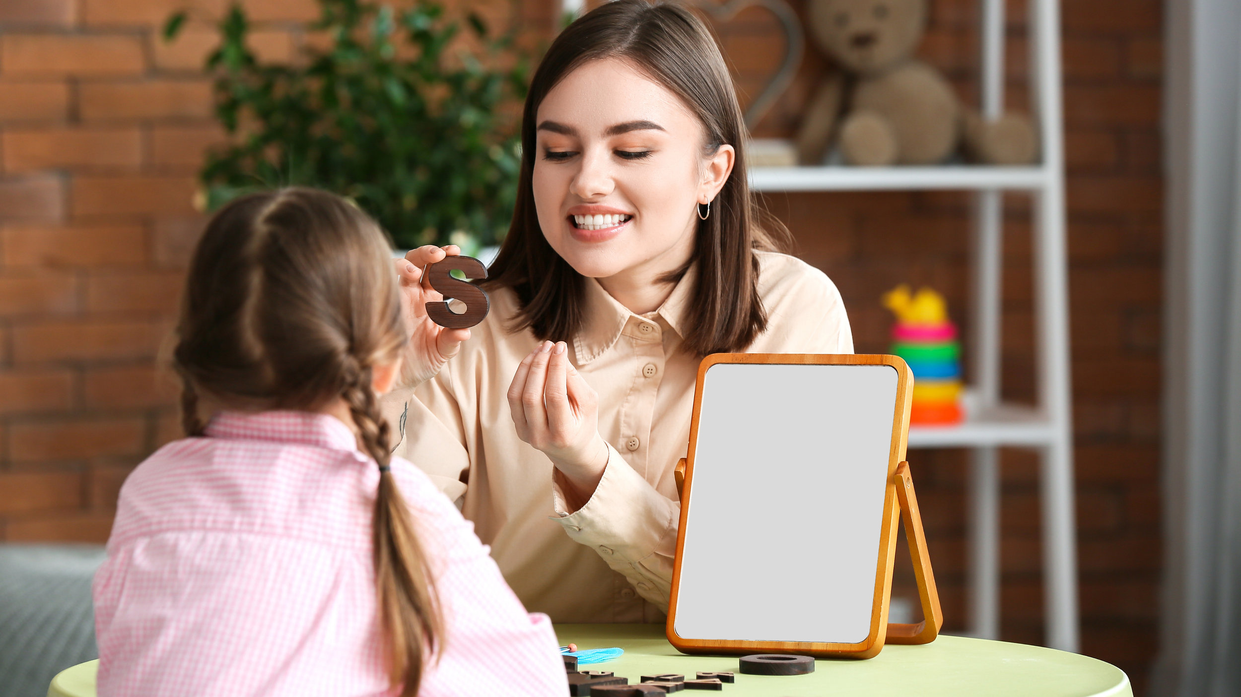 A speech pathologist teaching a young girl in a classroom, holding a letter S cut-out, with a mirror on the table reflecting her face, a brick wall background, and colorful objects on a shelf.