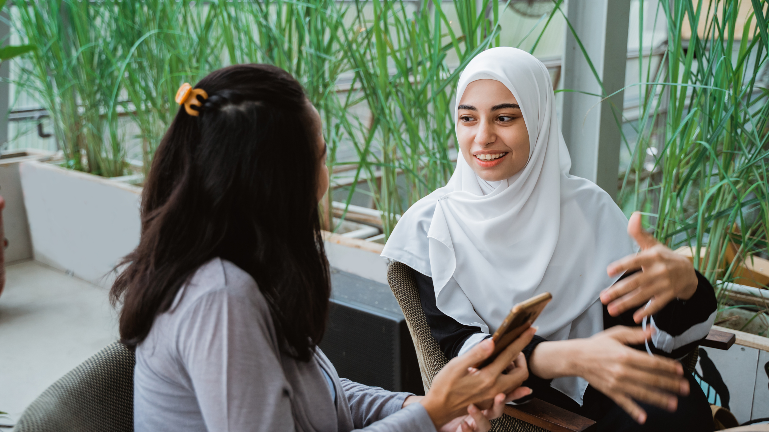 Psychology therapy session in a bright indoor setting with green plants in the background. One woman with dark hair and a hair clip holds a smartphone, while the woman with a white hijab who is the psychologist is smiling and gesturing.