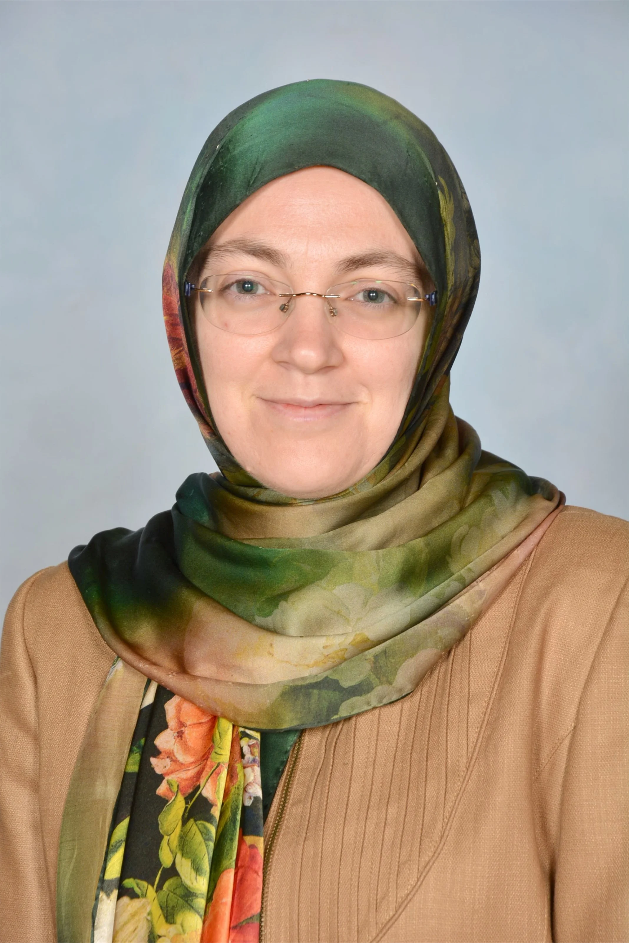A woman wearing glasses and a green, patterned headscarf, wearing a tan jacket, smiling softly against a plain, light background.