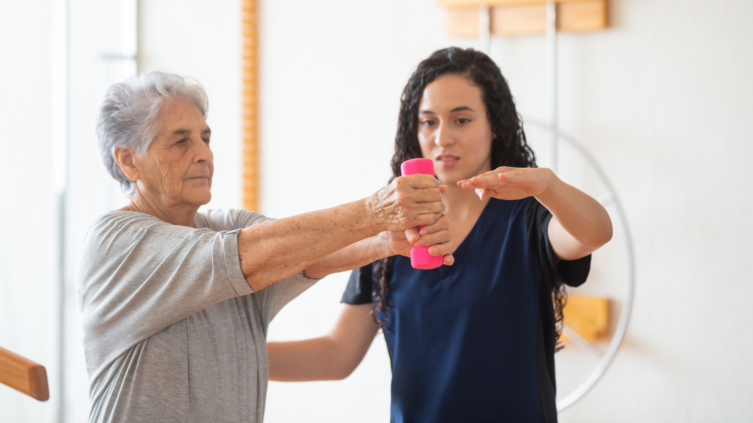An elderly woman and a young female instructor in a fitness or rehabilitation setting. The elderly woman is holding a small pink dumbbell while the instructor is helping her with an exercise. The instructor is providing support and guidance as the elderly woman performs a movement with her arm extended.