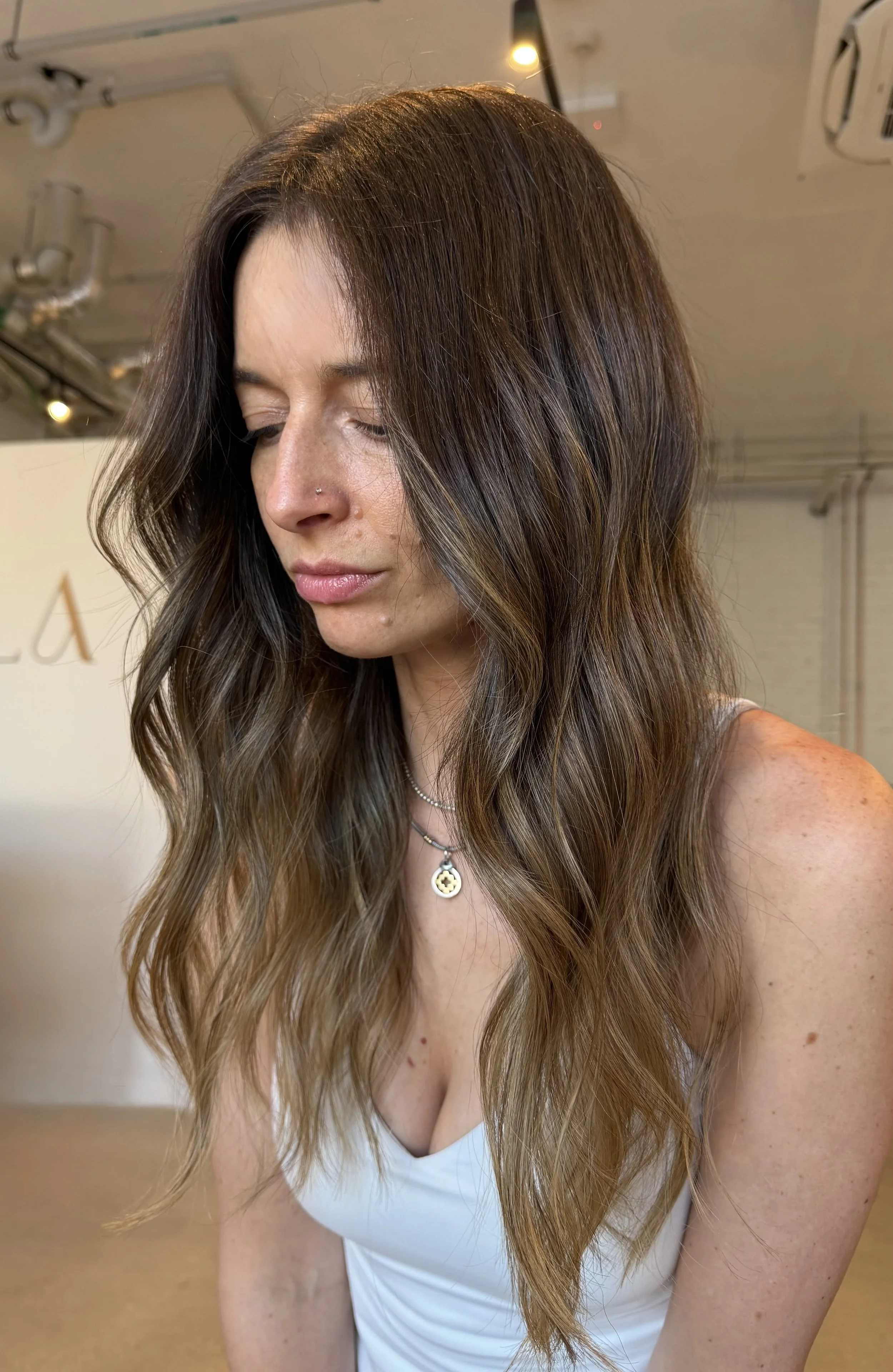A woman with long, wavy brown hair with subtle highlights,  in an indoor setting with warm lighting. Styling done with natural glam waves for a event