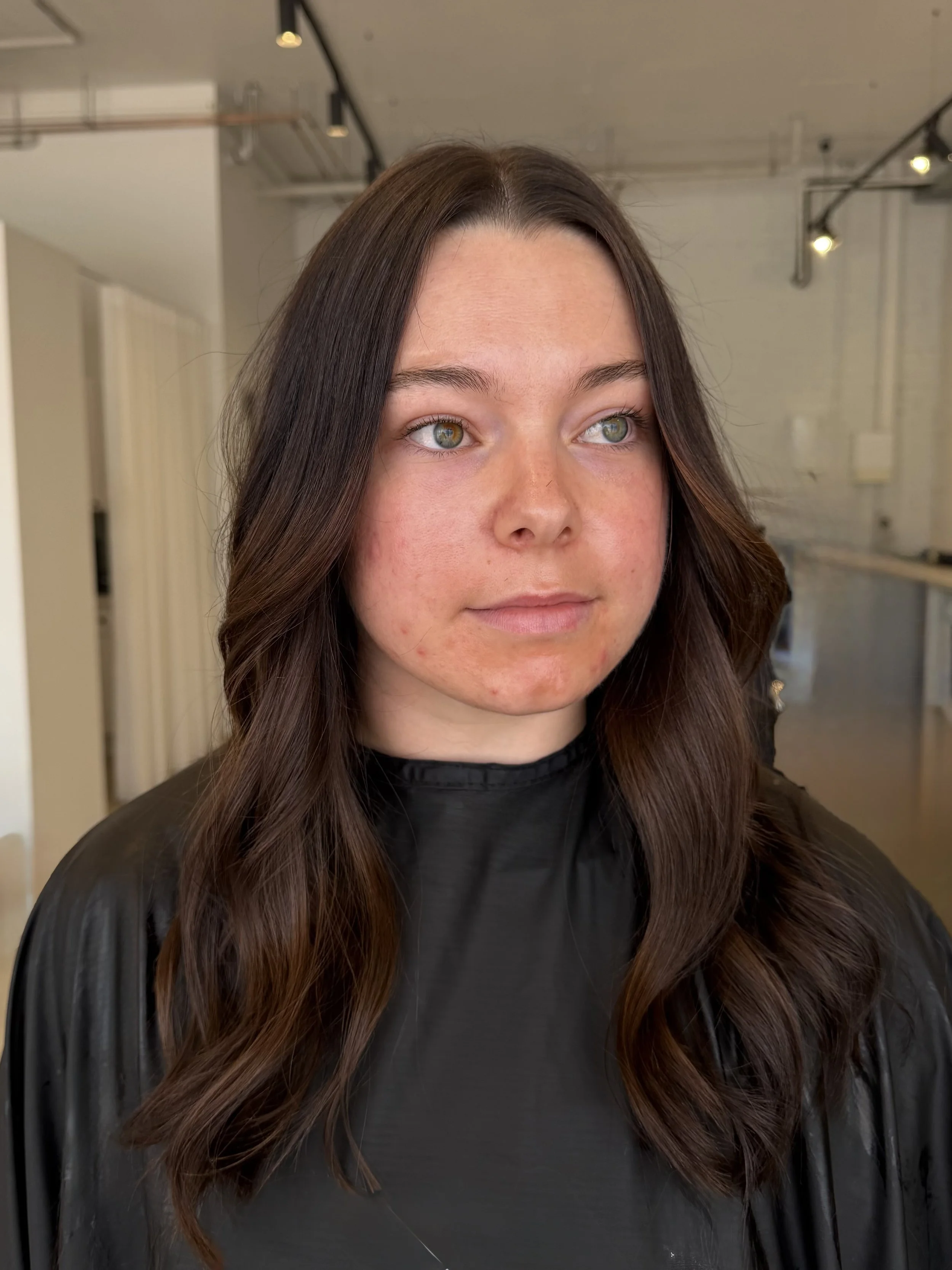 A woman with long wavy brown styled hair and fair skin, shiny and healthy brunette hair with treatment, bridal and bridges maid hair in Mount Lawley, appearing after a skincare or beauty treatment in a salon or studio, wearing a black cape.
