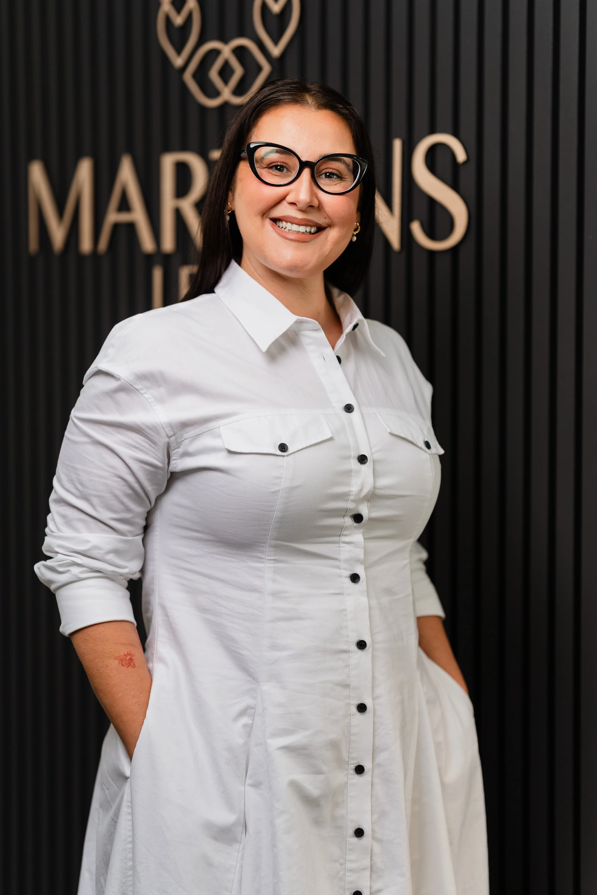 A woman in a white button-up shirt smiling with her hands in her pockets, standing in front of a black wall with gold lettering and designs.