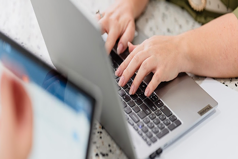 Close-up of a child's hands typing on a laptop keyboard, with an adult holding a smartphone nearby.