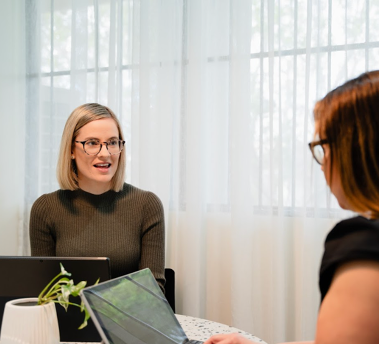 Two women having a discussion at a table with laptops, near a window with white curtains