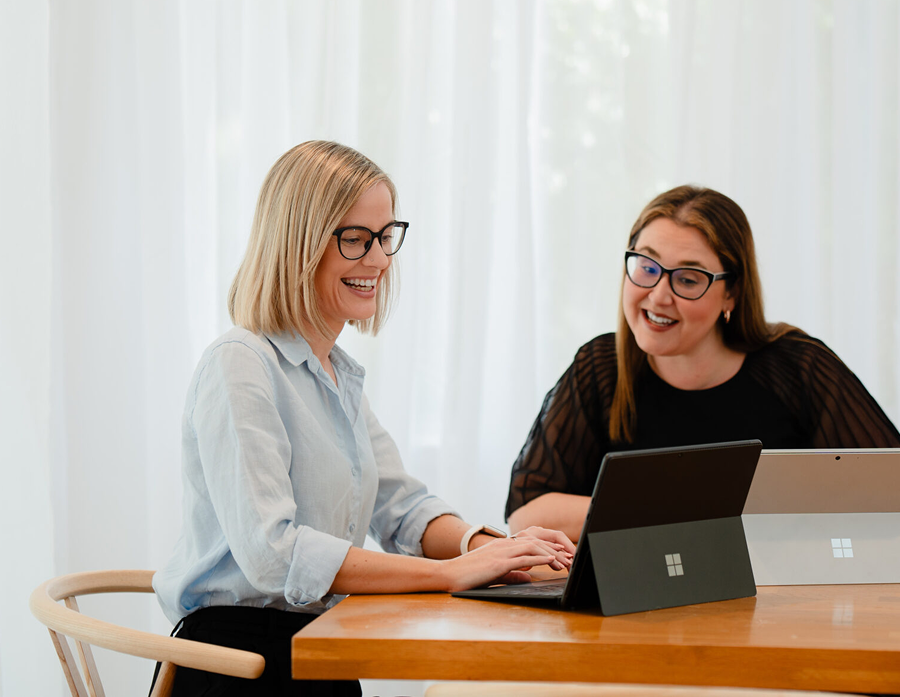 Two women with glasses working together on a laptop at a wooden table, smiling, in a bright room with white curtains.