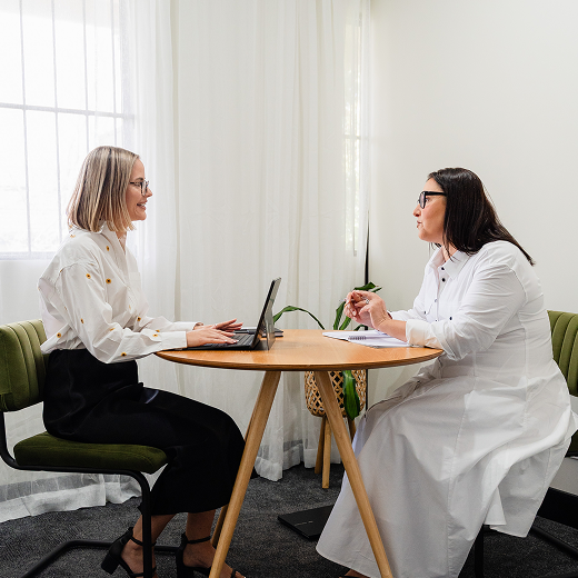 Two women sitting at a round wooden table, engaged in a conversation, one is using a laptop, the other is taking notes, in a bright room with large windows and white curtains.
