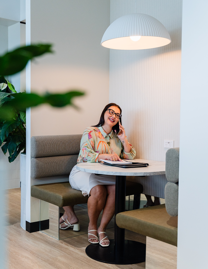 A woman with glasses and long dark hair sitting in a booth at a round table, talking on her phone and smiling, with a large white pendant light overhead and a green plant partially visible in the foreground.