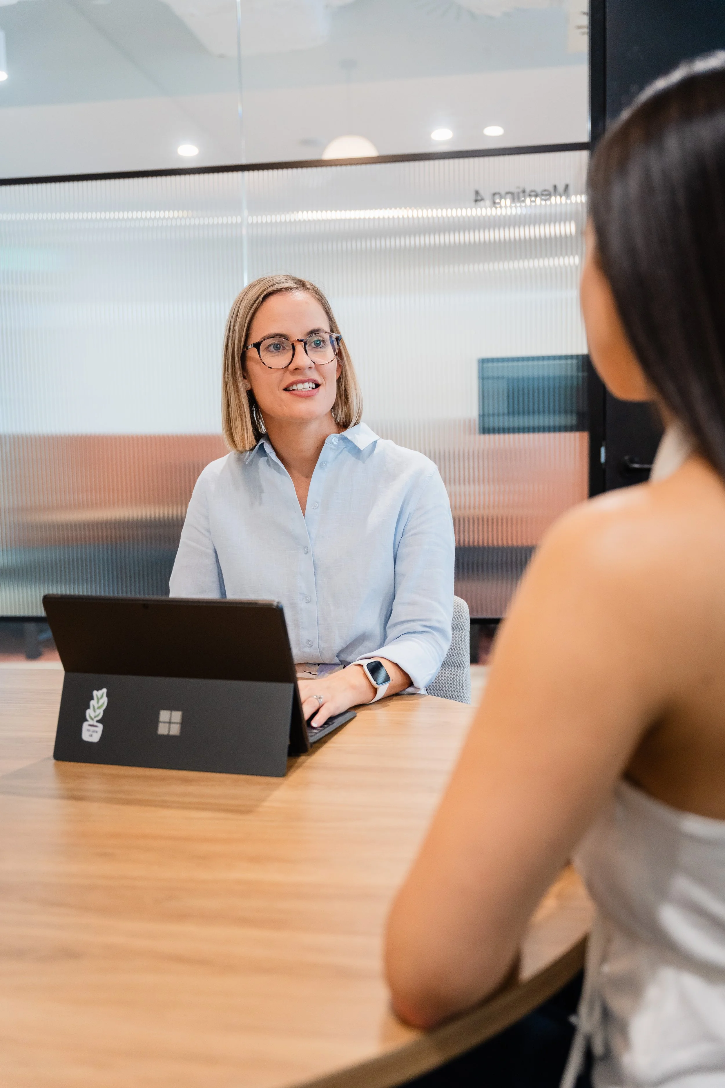 A woman with glasses and a light blue shirt sitting at a desk having a conversation with another woman in a white sleeveless top in a modern office environment.