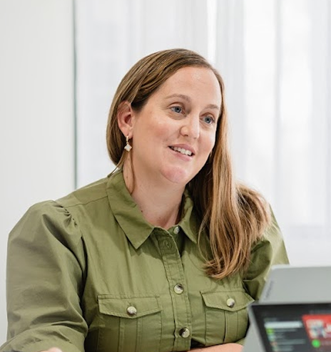 A woman with long, light brown hair wearing a green button-up shirt, sitting at a table with a laptop and tablet, smiling and looking to the side, in a bright room with white curtains.