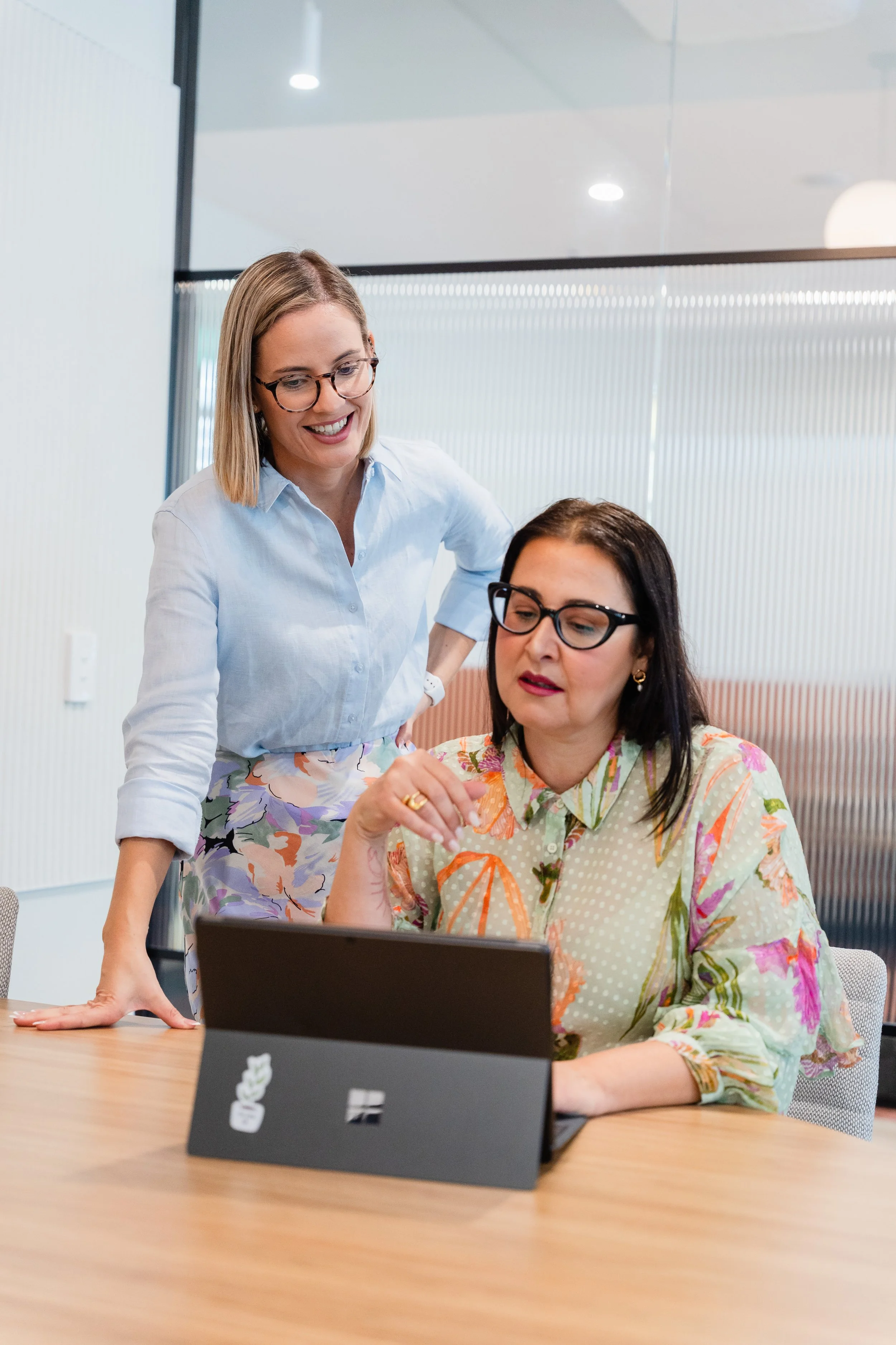 Two women working together in an office, one standing and the other sitting at a table with a tablet in front of her.