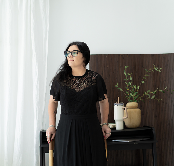 Woman with black hair and glasses standing indoors in front of a dark wooden panel with a potted plant and decorative items on a black table.
