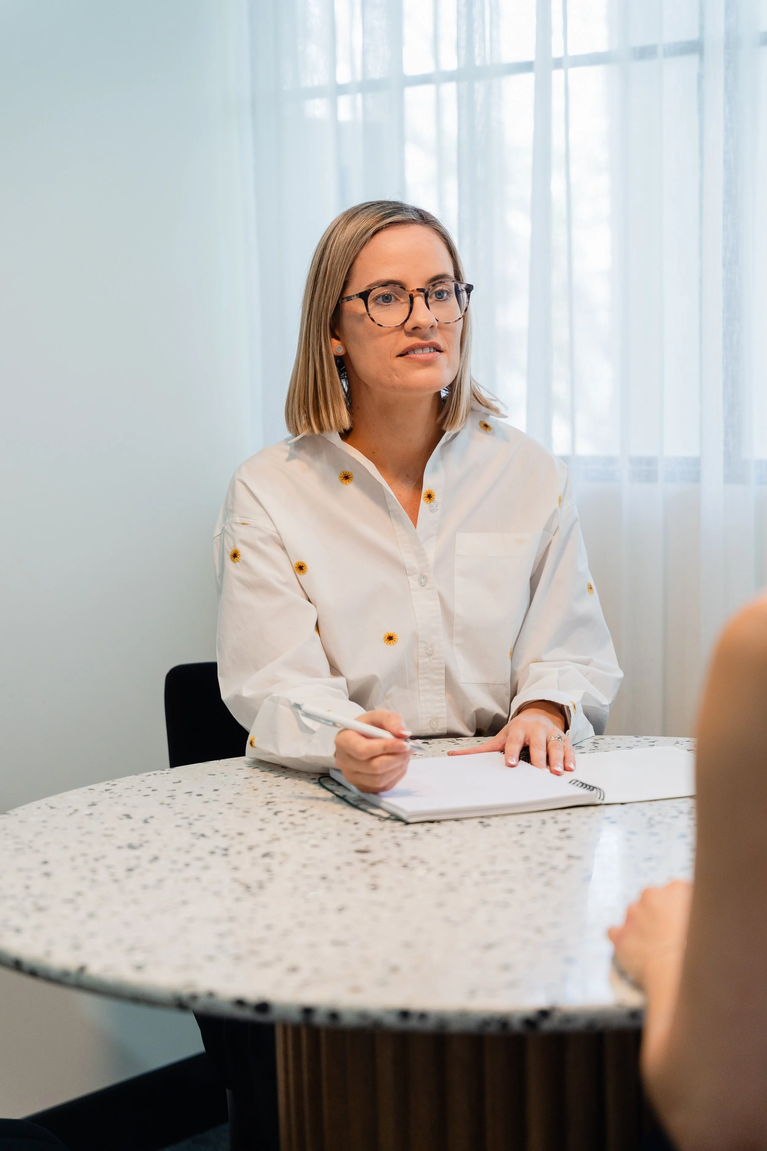 A woman with shoulder-length blonde hair, wearing glasses and a white shirt with small yellow flower patterns, is sitting at a round, speckled table. She is holding a pen and has a notebook open in front of her. There are sheer curtains and a window in the background.