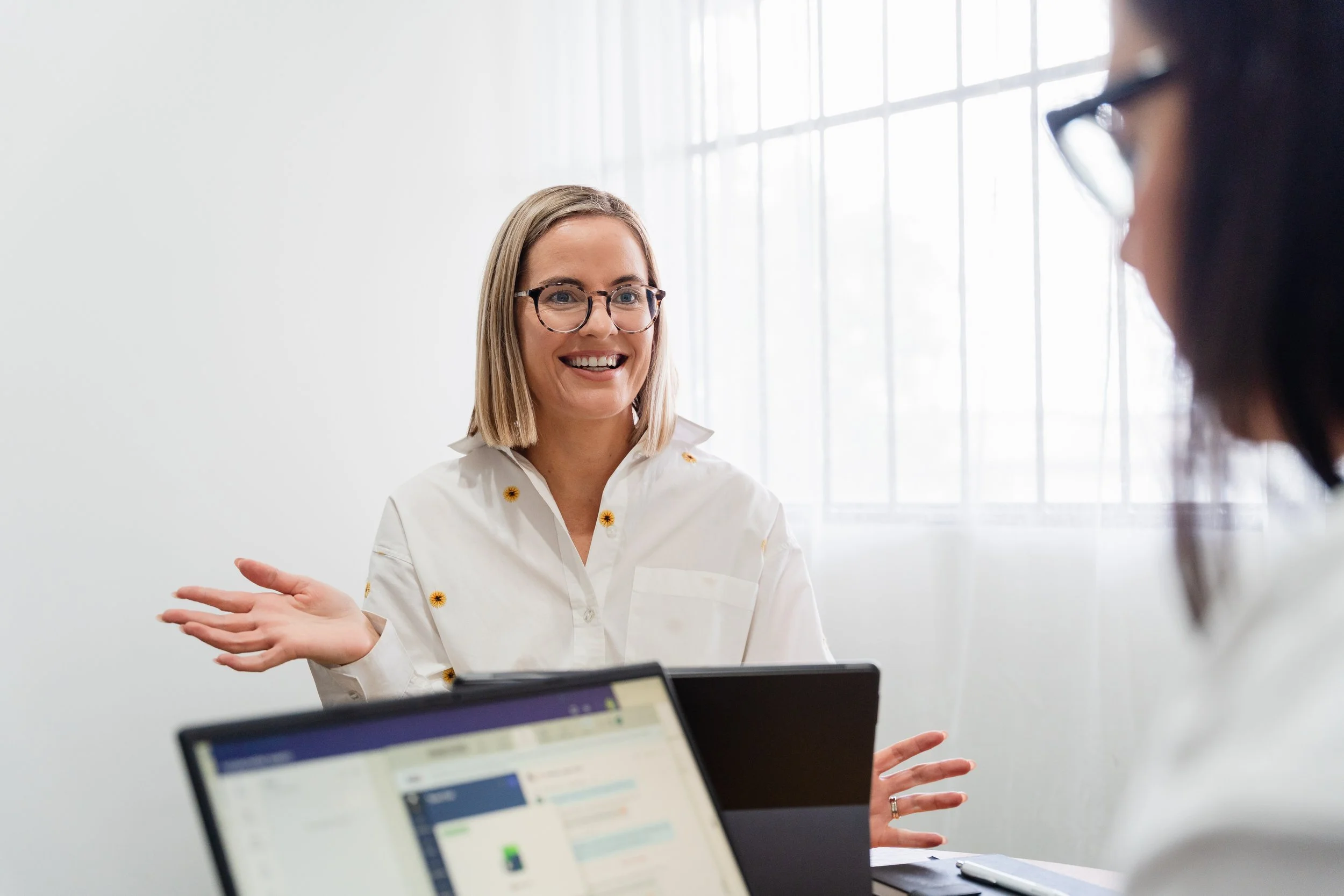 Two women in an office meeting, one smiling and gesturing, the other partially visible with a laptop in front.