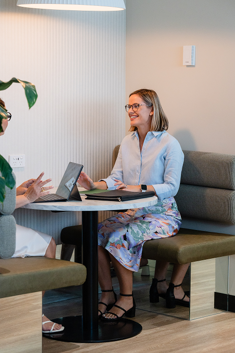 Two women having a conversation at a table in a modern office or cafe, one woman with glasses and a light blue shirt smiling, the other woman's face is not visible, a laptop and notebook on the table.