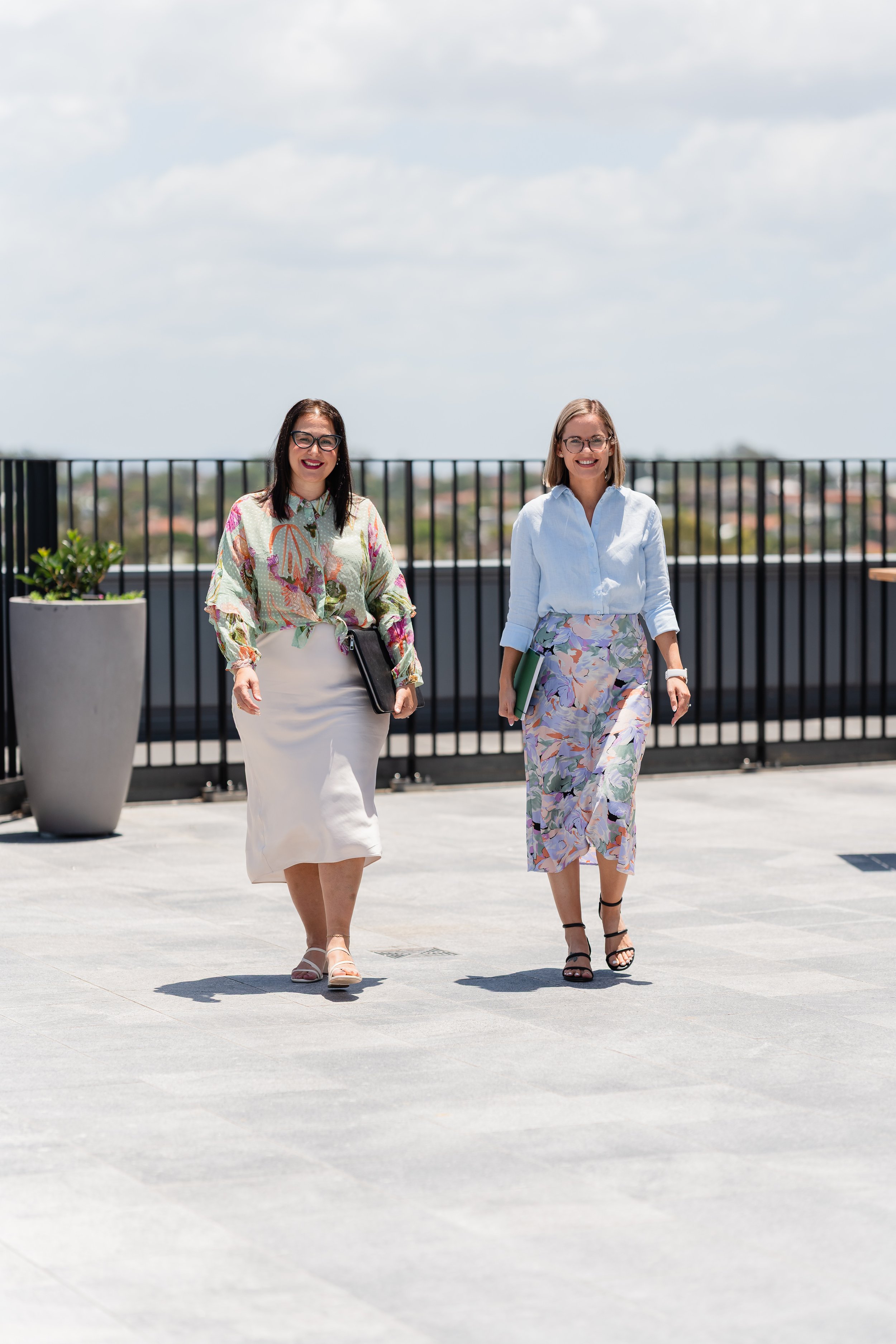 Two women walking outdoors on a sunny day, holding notebooks, with a black metal fence and a cityscape in the background.
