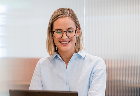 Woman with glasses smiling while looking at a laptop in an office setting.