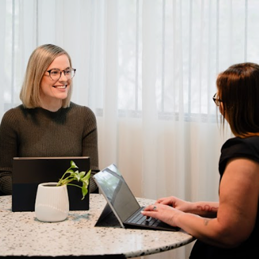 Two women sitting at a table having a conversation, one with blonde hair and glasses, the other with dark hair and glasses, with laptops and a potted plant on the table.