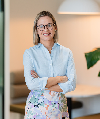 A smiling woman with glasses and blonde hair standing indoors, wearing a light blue blouse and a floral skirt, with her arms crossed.