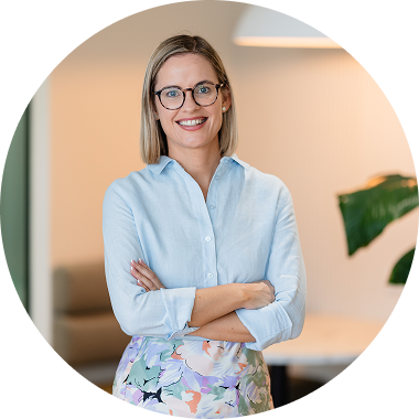A smiling woman with glasses, wearing a blue blouse and patterned skirt, standing in an indoor setting with a blurred background, including a large green plant.