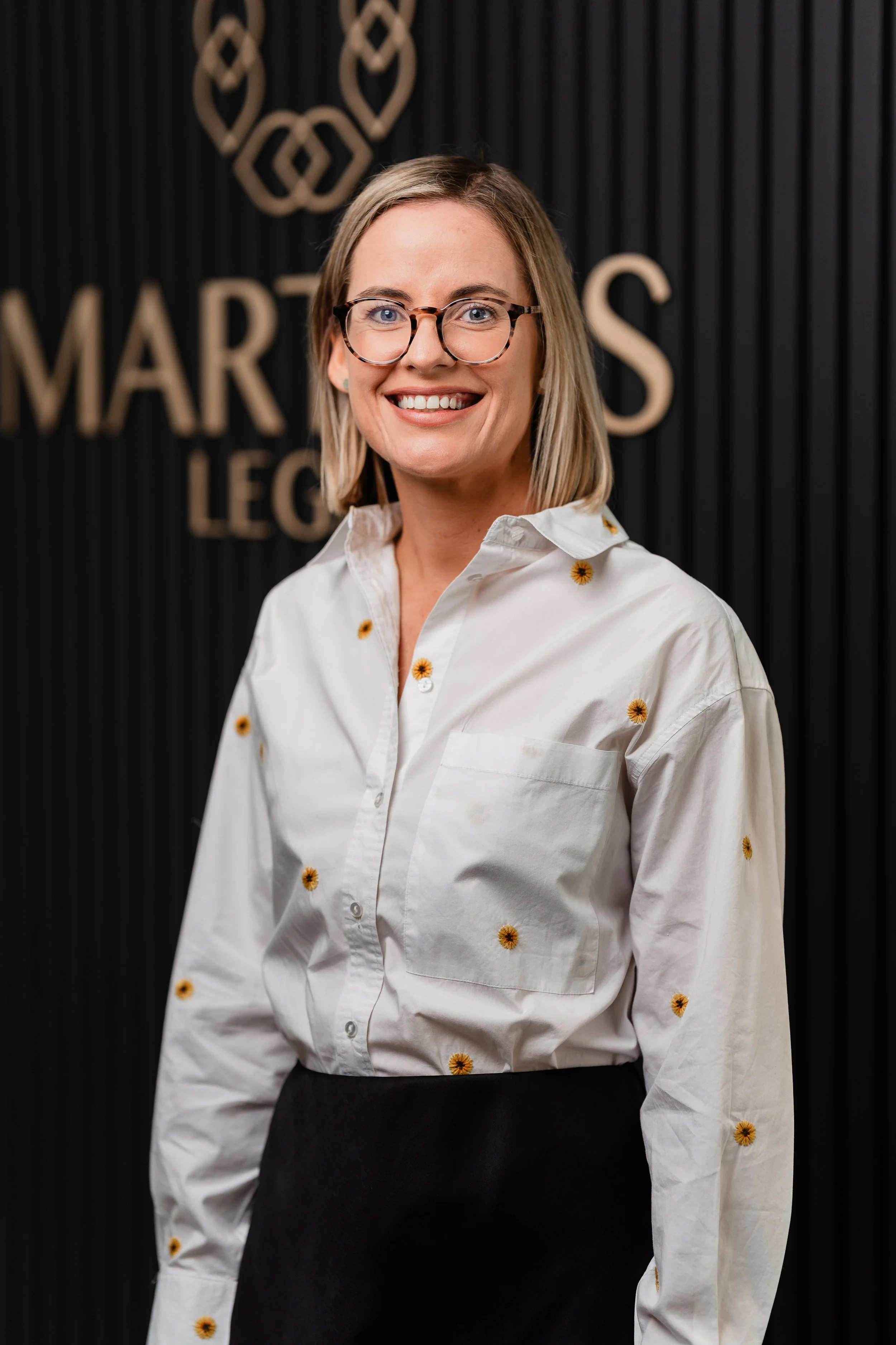A smiling woman with shoulder-length blonde hair, wearing glasses, a white shirt with small embroidered flowers, and a black skirt, standing indoors against a black textured wall with gold lettering and design.