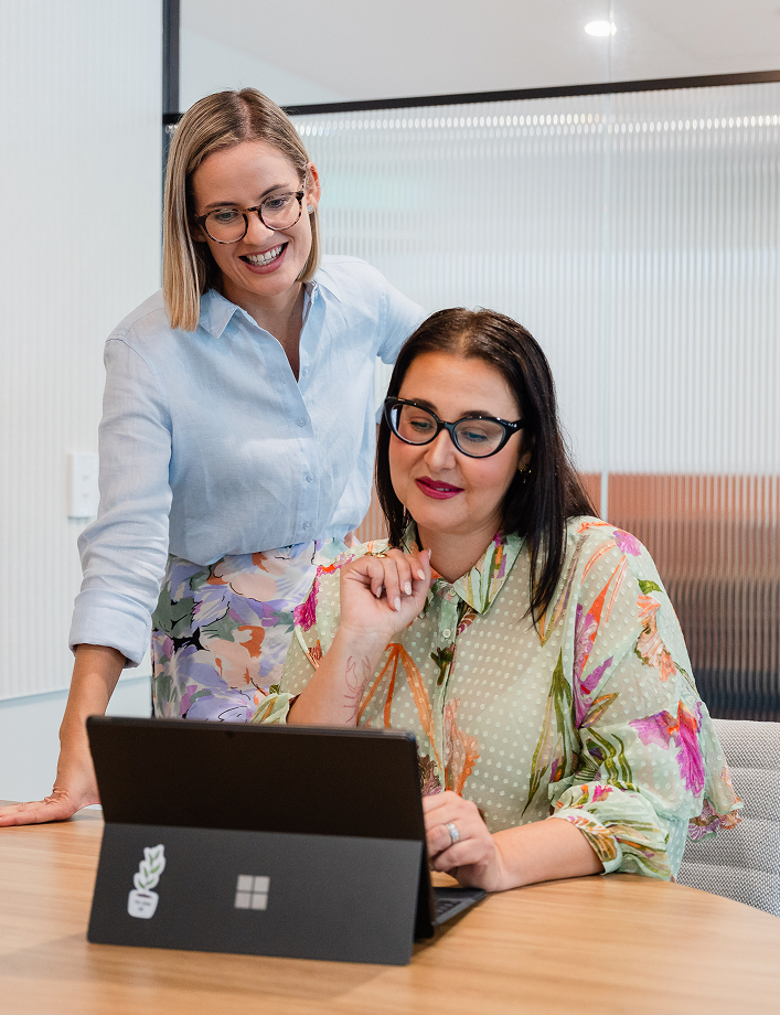 Two women working together on a laptop in an office, one sitting and the other standing beside her.