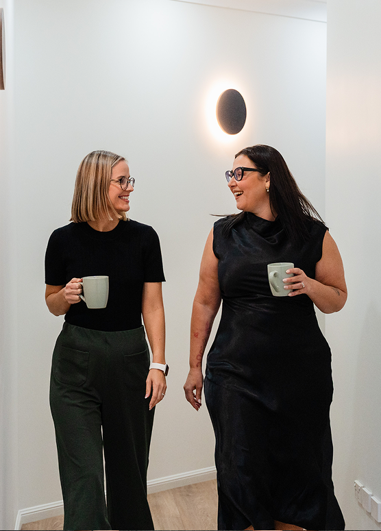 Two women smiling and walking together in an indoor setting, each holding a coffee mug.