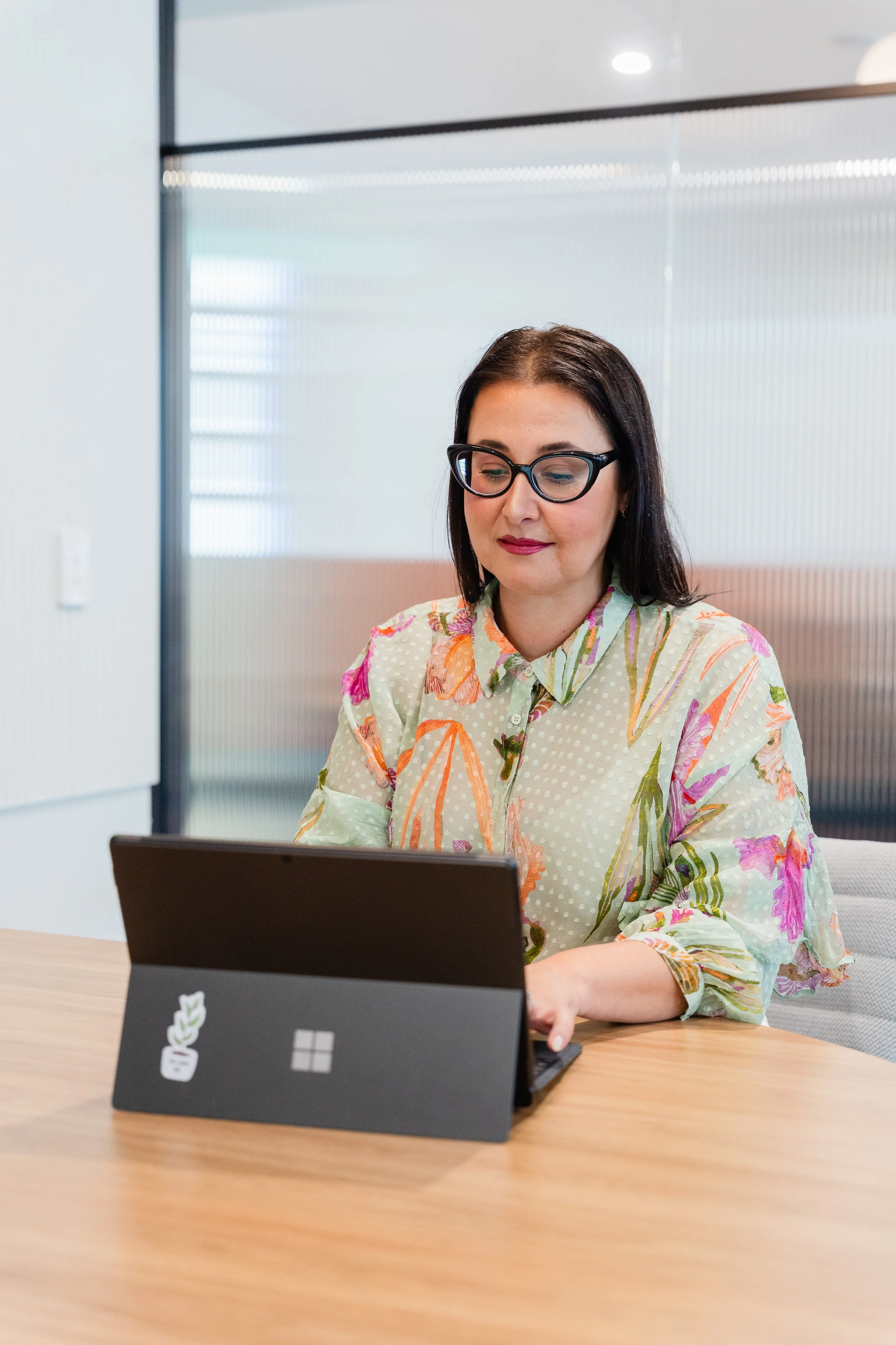 A woman with dark hair, wearing glasses and a floral blouse, sitting at a wooden table, working on a Microsoft Surface tablet in a modern office with frosted glass partitions.