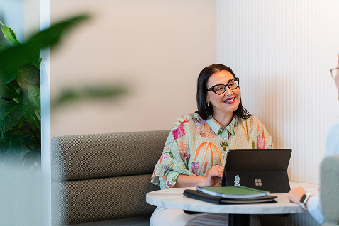 Woman with glasses smiling at a laptop during a conversation in an office.