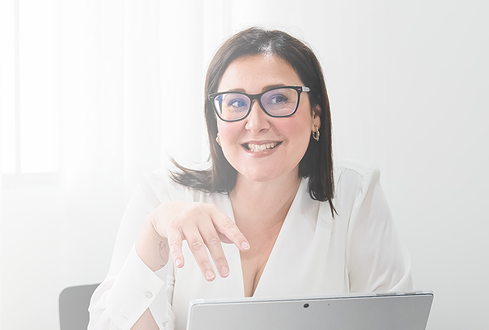 A woman with dark hair, glasses, and earrings smiling at the camera, sitting in front of a laptop in a bright room.