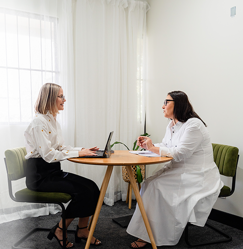 Two women sitting at a small round table in an office, engaging in a discussion. One woman has blonde hair, glasses, and is using a laptop. The other woman has dark hair, glasses, and is wearing a white lab coat. The room has white walls, a window with white curtains, and green chairs.