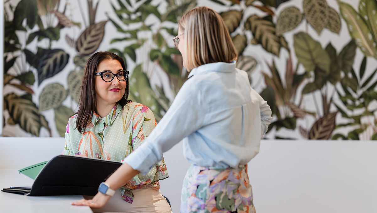 Two women having a conversation in an office with a tropical leaf-patterned wall background.