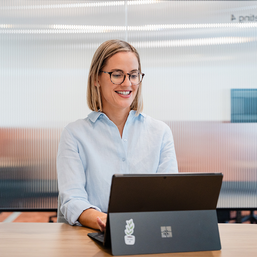 A woman with glasses sitting at a desk, working on a Microsoft Surface laptop in an office setting.