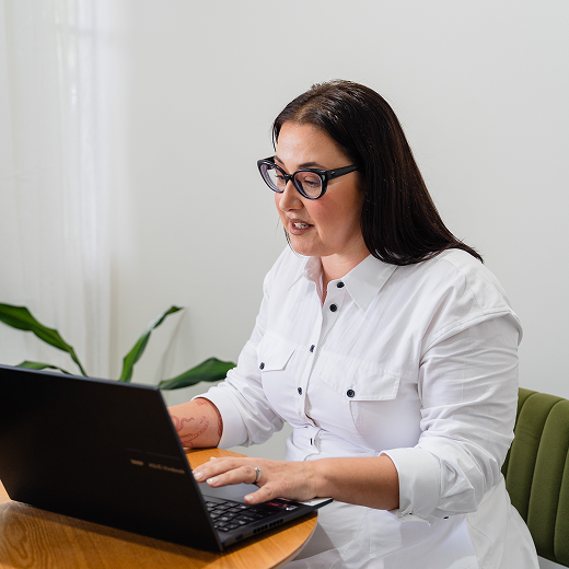 Woman with dark hair and glasses working on a laptop at a wooden table in a bright room with white walls.