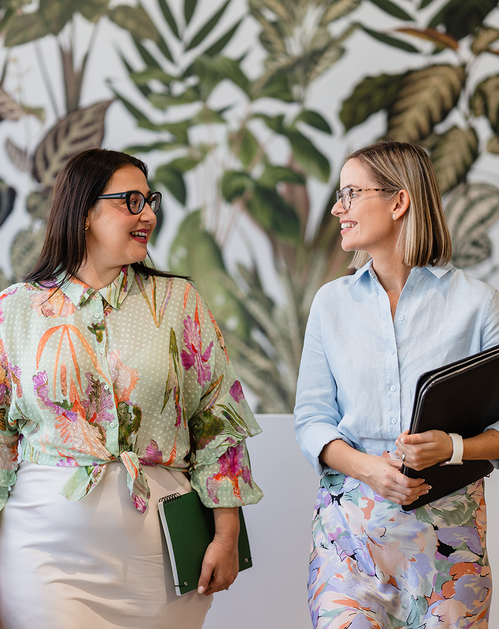 Two women having a conversation indoors, both holding notebooks, with a leafy plant mural in the background.