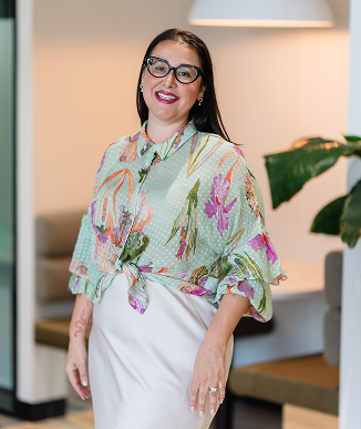 Woman with glasses and long dark hair wearing a colorful floral blouse and white skirt, smiling indoors with a plant and seating in the background.