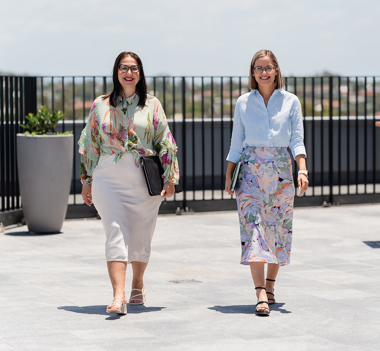 Two women walking on a rooftop terrace with black railing, city view in the background, wearing stylish summer outfits and sunglasses.