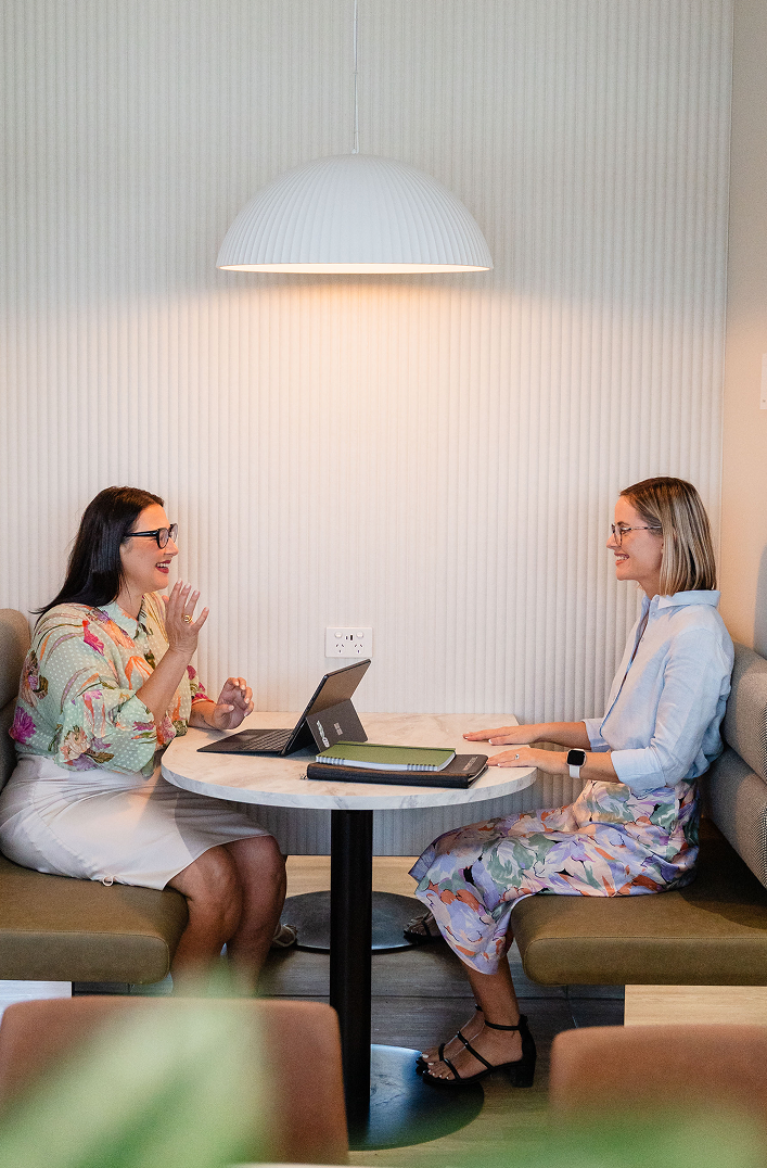 Two women sitting at a round table in a cafe, engaging in a friendly conversation. One woman is wearing a floral blouse and white skirt, the other is in a light blue blouse and floral skirt. There are notebooks and a tablet on the table, and a large white pendant light hangs above.