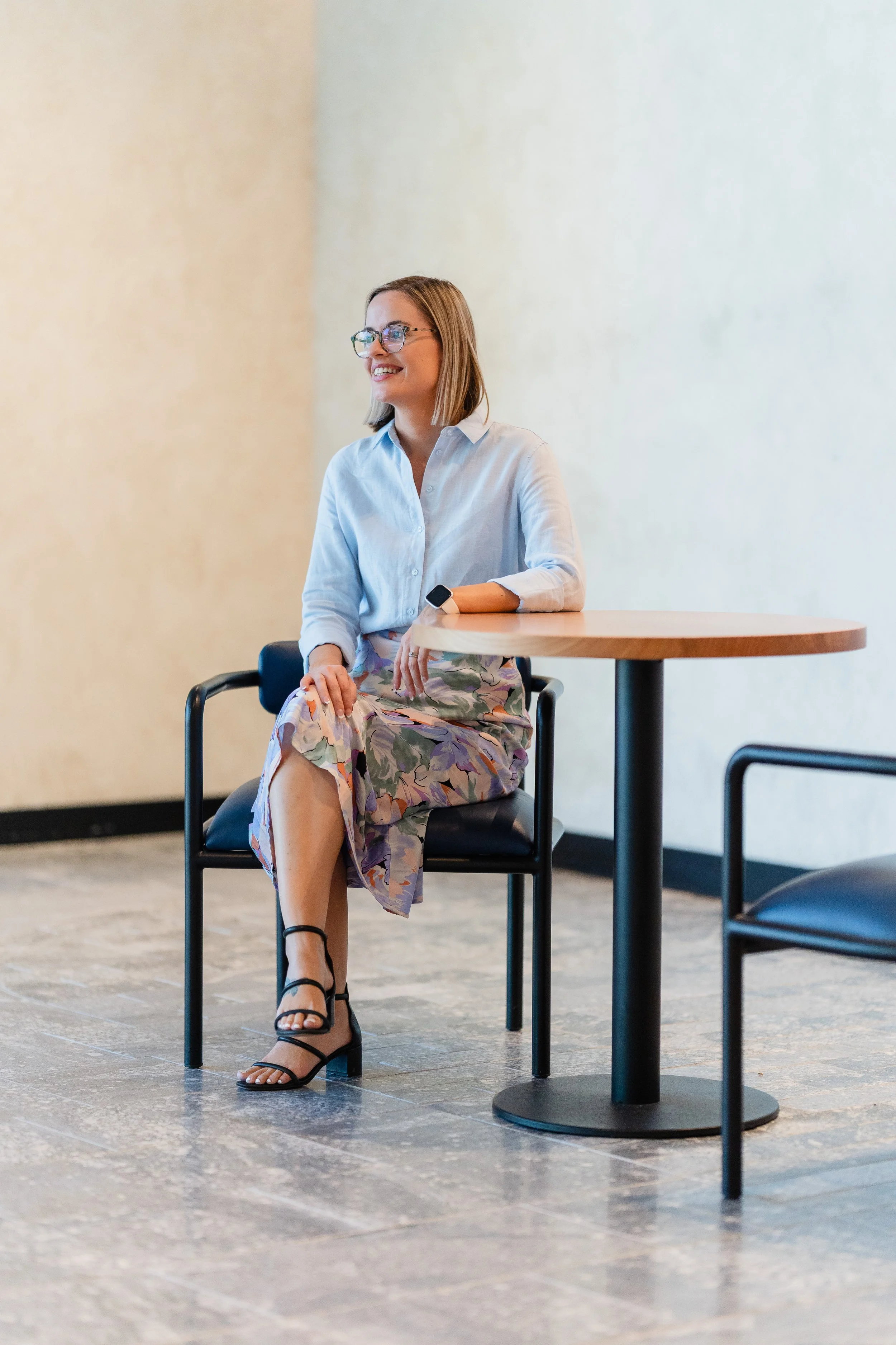 A woman with glasses in a white blouse and floral skirt sitting at a round table in a modern indoor setting.
