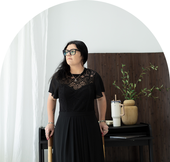 Woman with glasses and black dress standing in a room with a plant and decorative objects on a table.